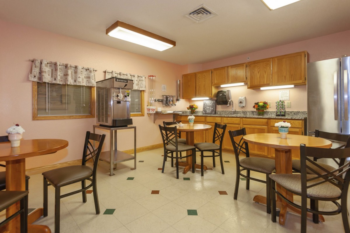 A cozy dining area with wooden tables and chairs, featuring a kitchen setup with cabinets and overhead lighting.