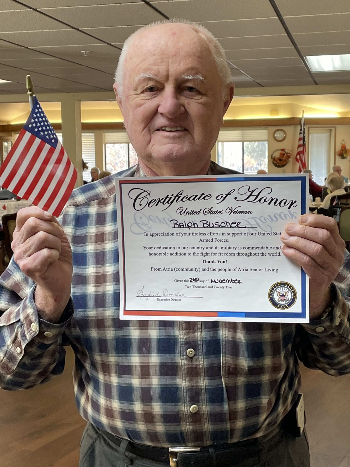 A man holding a Certificate of Honor and an American flag indoors.