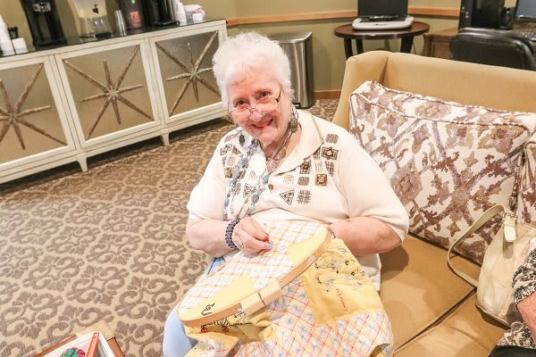An elderly woman is sitting on a couch and smiling while working on a sewing project.
