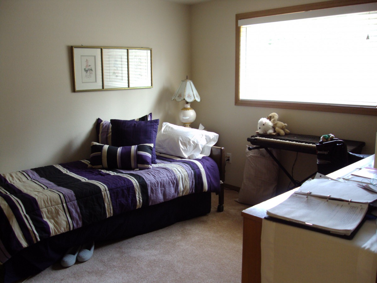 A cozy bedroom with a single bed featuring striped bedding, decorative pillows, and a lamp by the window. A desk is positioned in front of the window.