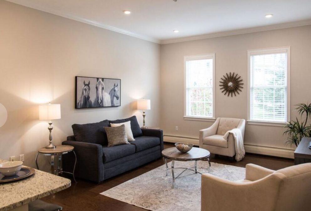 A modern living room with neutral tones, featuring a dark gray sofa, two armchairs, and a central coffee table. The space is accentuated by wall art and a sunburst mirror.