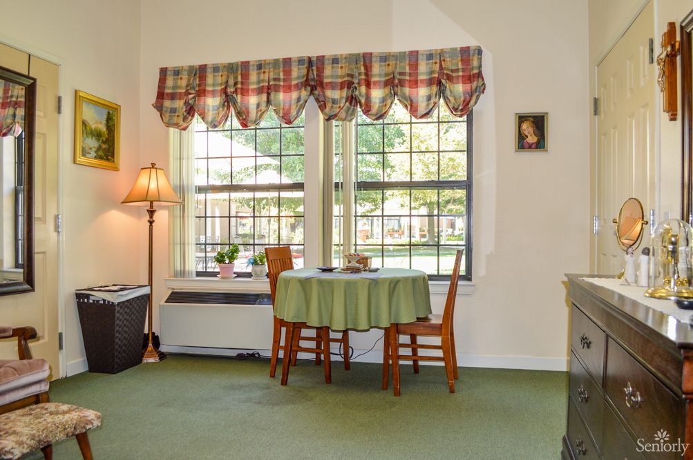 A cozy dining area featuring a round table and two wooden chairs by large windows with plaid curtains.