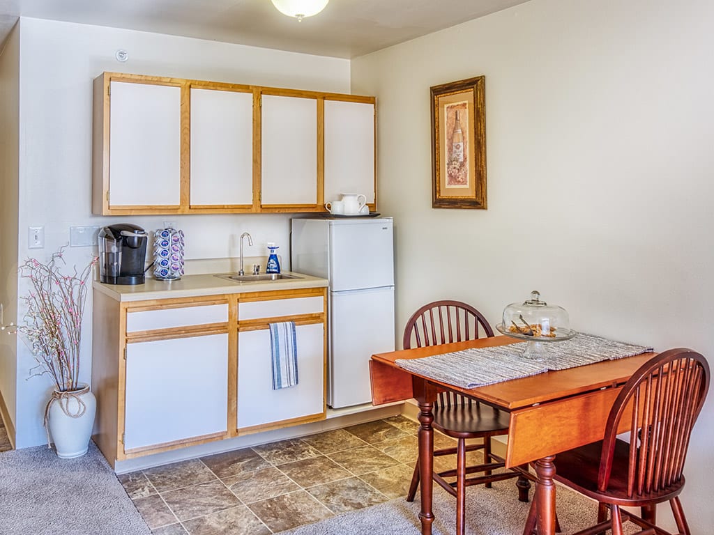 A compact kitchen and dining area featuring a small table and chairs, cabinets, a fridge, and a coffee maker.