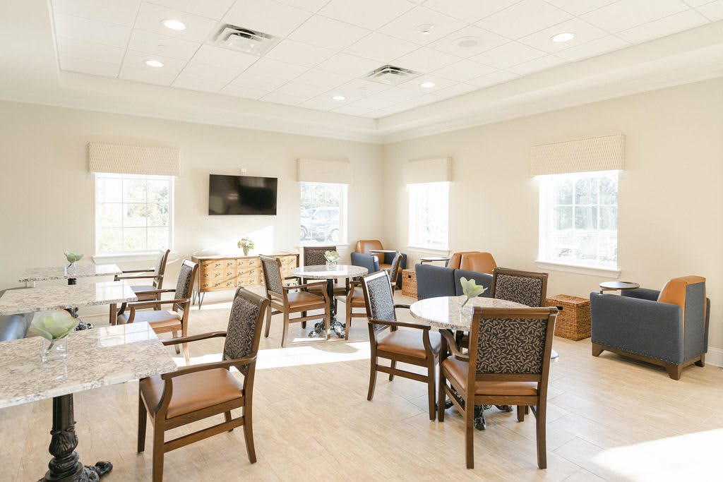 A bright and airy dining area with multiple tables and chairs, featuring a wall-mounted television and large windows.