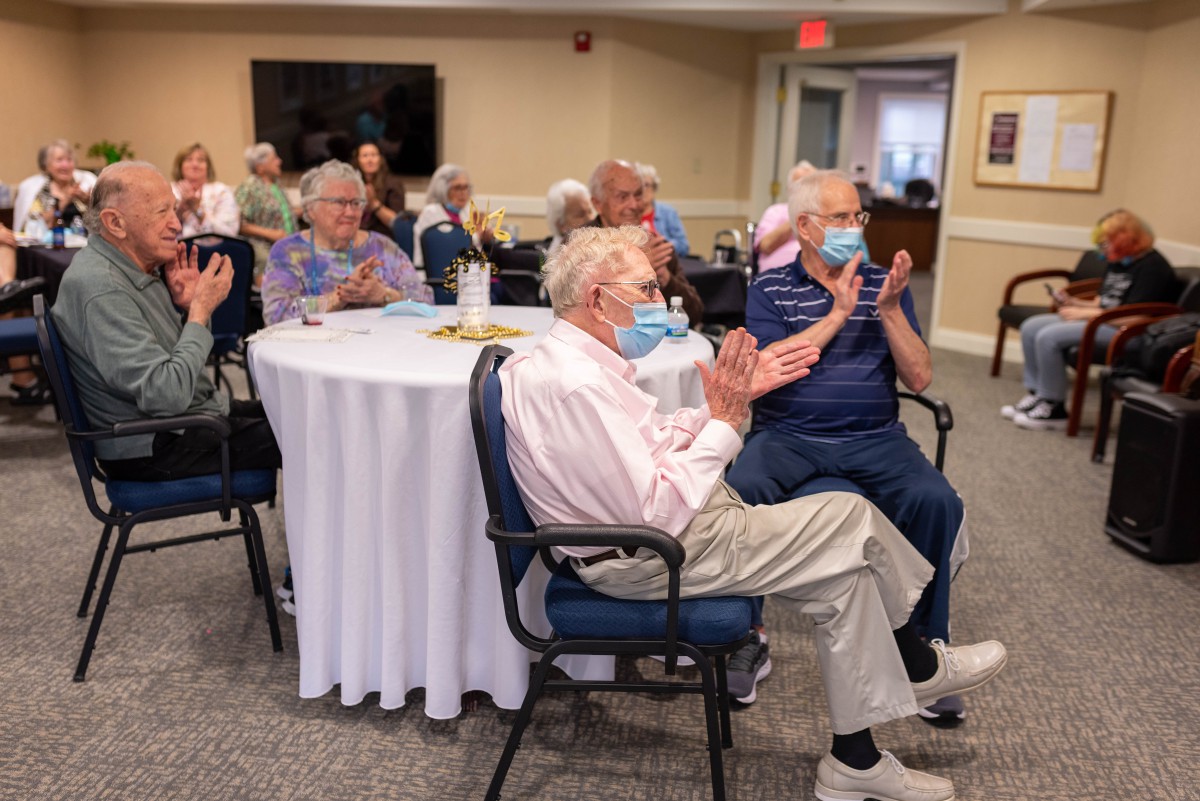 A group of seniors seated indoors, clapping and wearing masks during a community gathering.