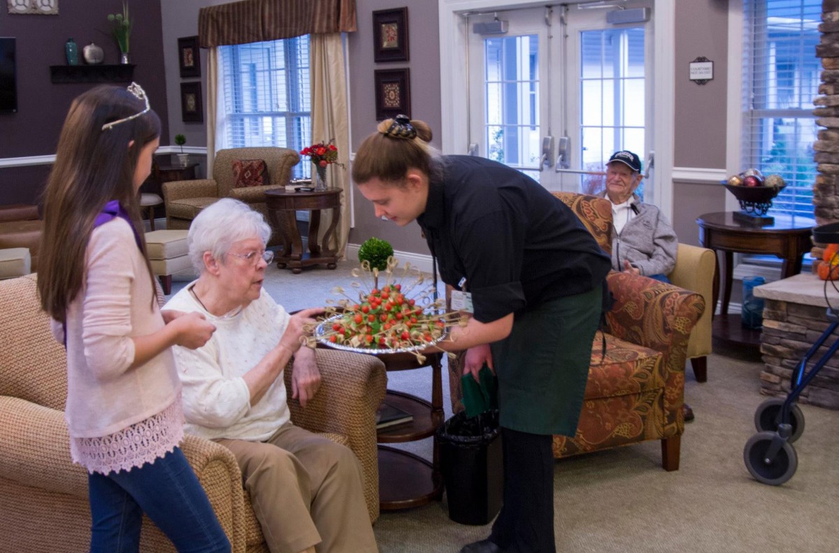 A caregiver serves food to an elderly resident in a nursing home, illustrating community and care.
