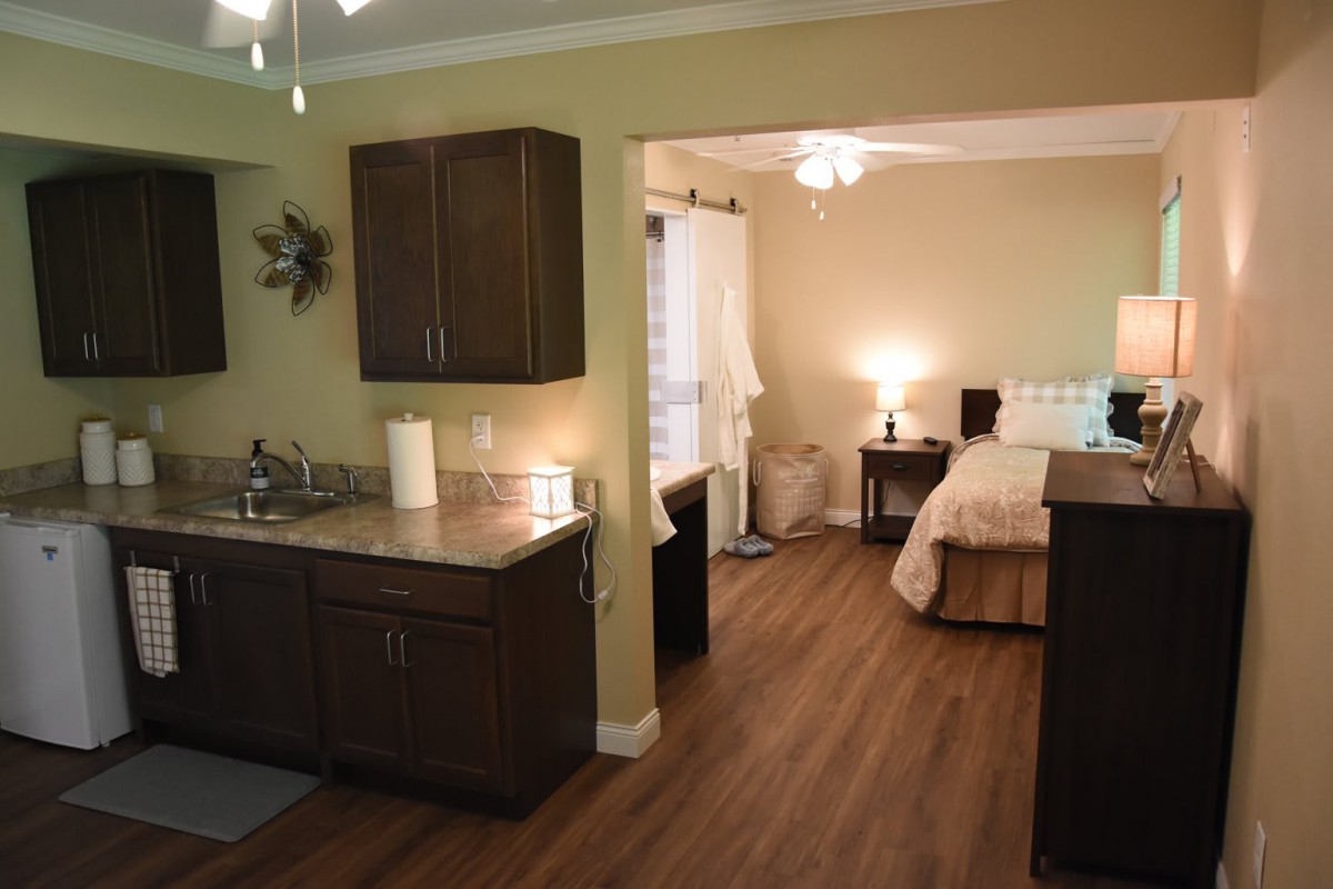 A cozy studio apartment with a kitchenette, wood flooring, and a well-lit bedroom area.