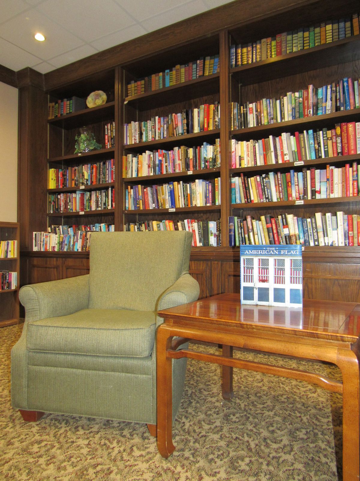A cozy reading area featuring a green armchair and a wooden table in front of a large bookshelf filled with books.