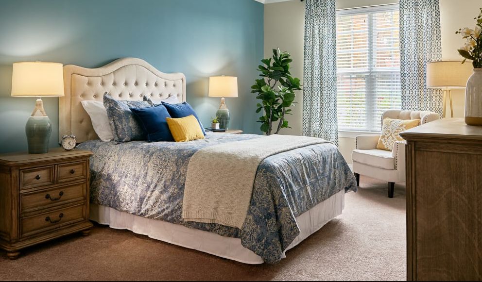 A cozy bedroom with a stylish bed, soft lighting from a lamp, and a large window letting in natural light.