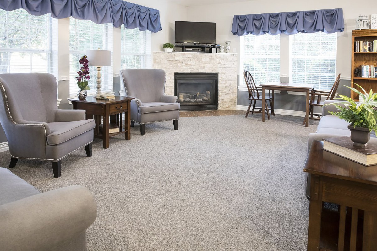 A cozy living room featuring armchairs, a fireplace, and a television, with large windows adorned by blue curtains.