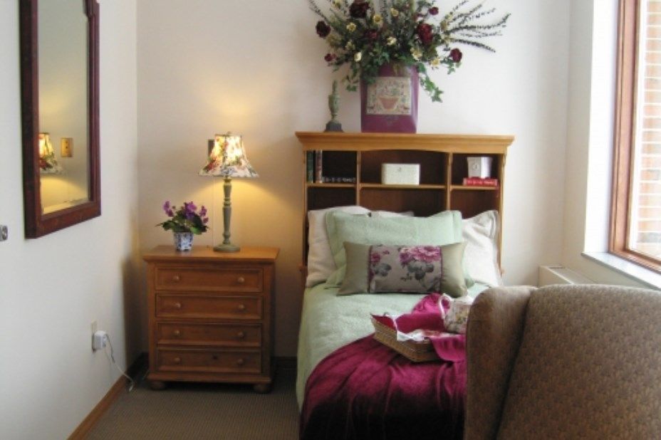 A cozy bedroom featuring a single bed with floral bedding, a wooden headboard, and a matching nightstand adorned with a lamp and vase of flowers.