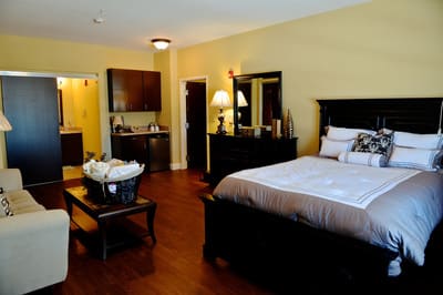 A cozy bedroom with wooden flooring, featuring a large bed, dresser, and ambient lighting.