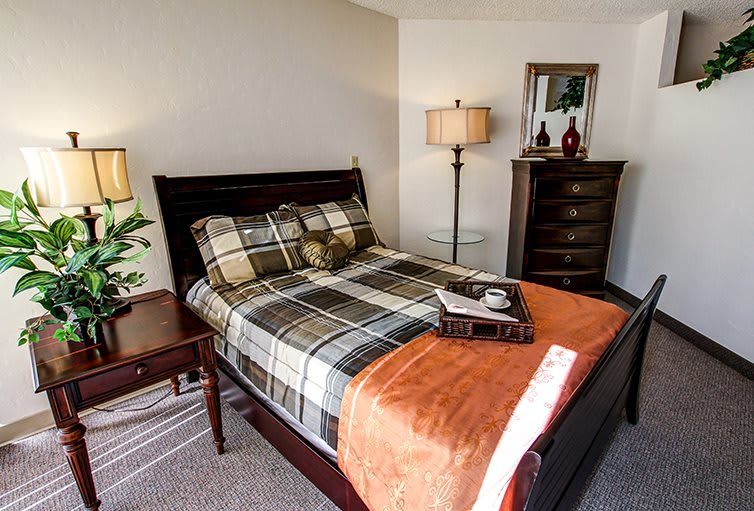 A cozy and well-decorated bedroom featuring a plaid bedspread, a warm orange throw blanket, wooden furniture, and a lamp.
