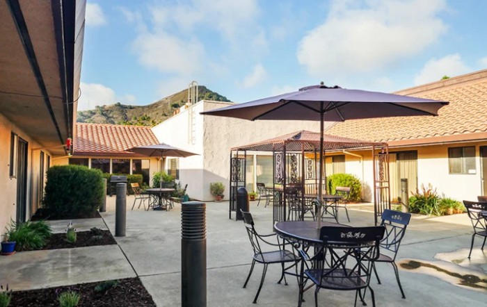 A patio area featuring several tables and chairs with umbrellas, set against a building and a blue sky.