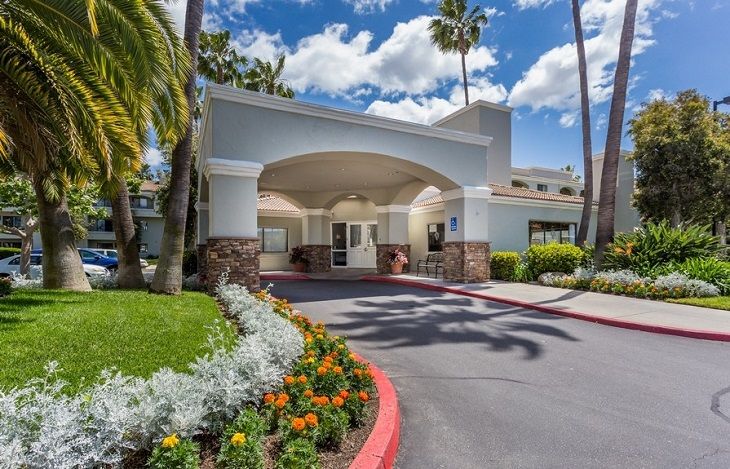 A hotel entrance with well-maintained landscaping, featuring palm trees and a parking area.