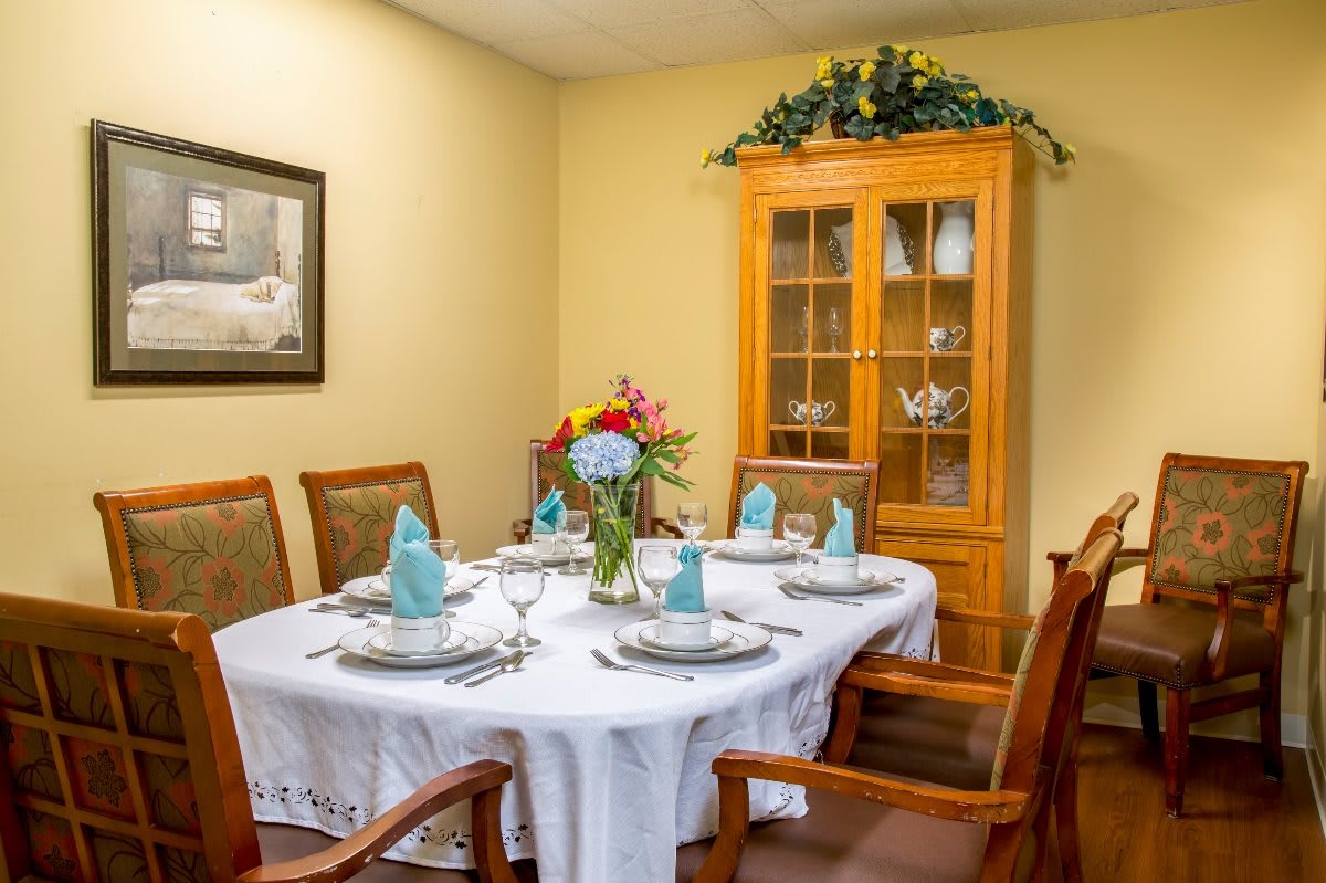 A neatly arranged dining room with a table set for six, featuring a floral centerpiece and an elegant china cabinet in the background.