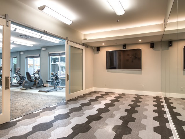 A modern exercise room with geometric floor tiles, featuring gym equipment and a glass partition.