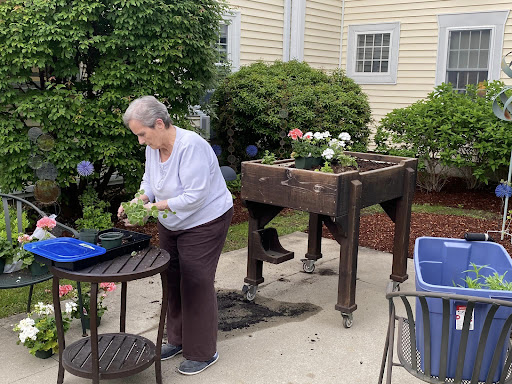 An elderly woman tending to her garden at a potting table, surrounded by blooming flowers and lush greenery.