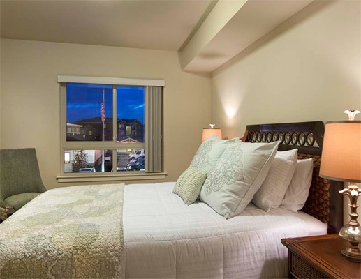 A cozy bedroom with a large bed, adorned with pillows and a bedside lamp, offering a nighttime city view from the window.