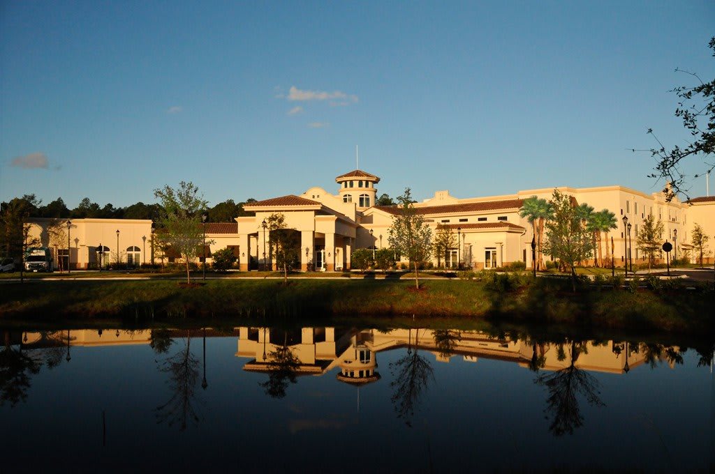 A large building with Mediterranean-style architecture is beautifully reflected in a calm lake during sunset.