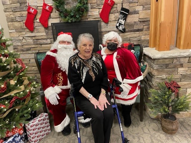 A festive holiday scene with two people dressed as Santa Claus posing beside an elderly woman, surrounded by Christmas decorations.