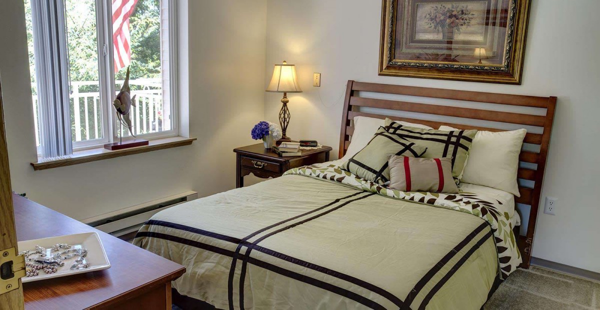 A cozy bedroom with a neatly made bed, bedside lamp, and a window providing natural light.