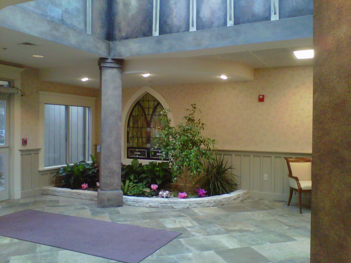 An indoor lobby with plants and seating, featuring a column and decorative arch window.