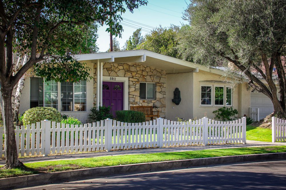 A quaint single-story house with a white picket fence and surrounded by trees.