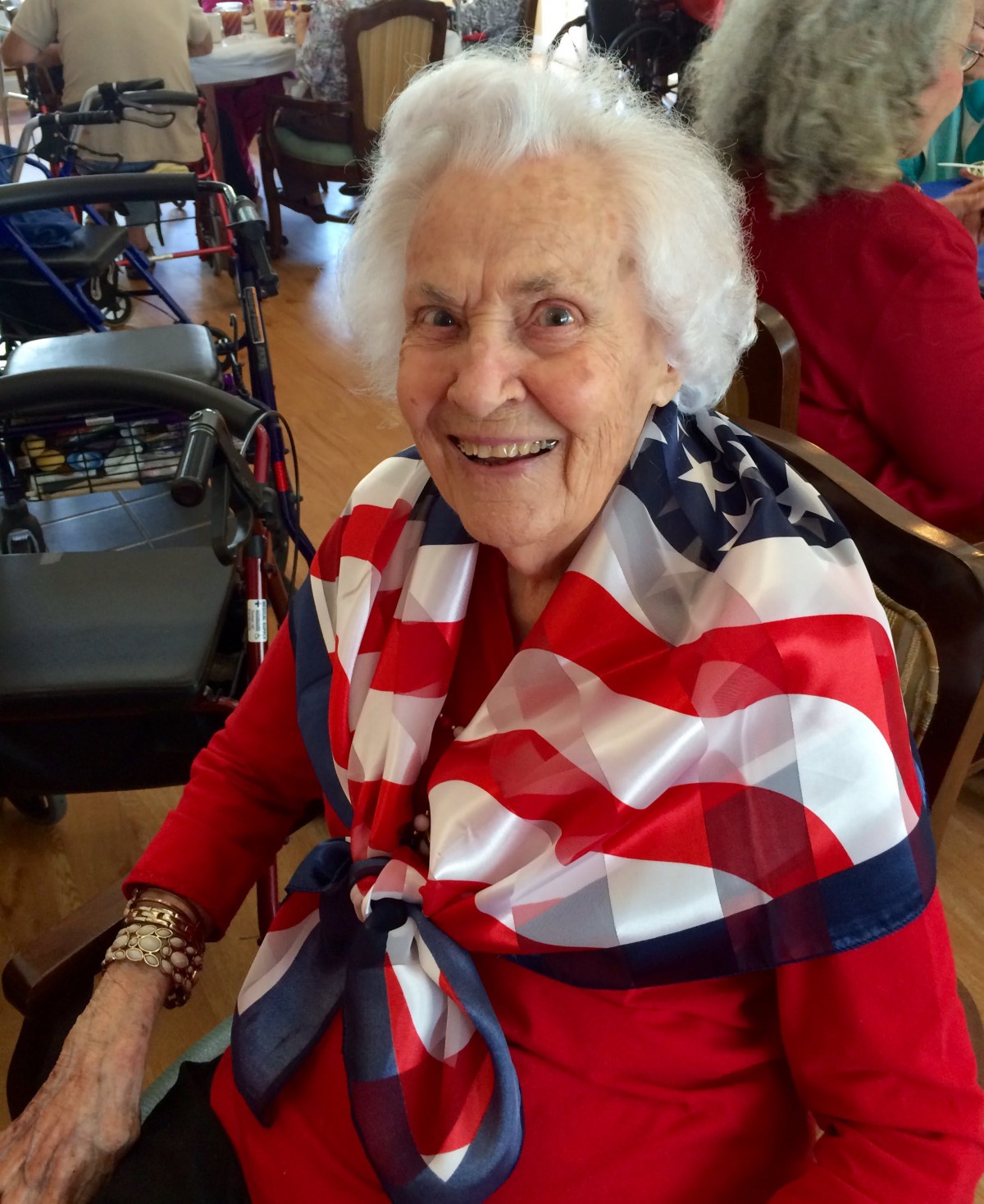 An elderly woman wearing a scarf with the American flag pattern and a red outfit, smiling warmly.
