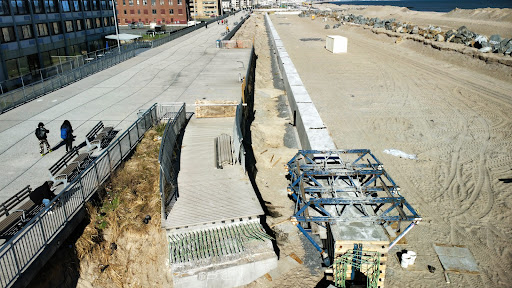 An aerial view of a construction site along the coastline, featuring a newly built path and materials ready for installation.