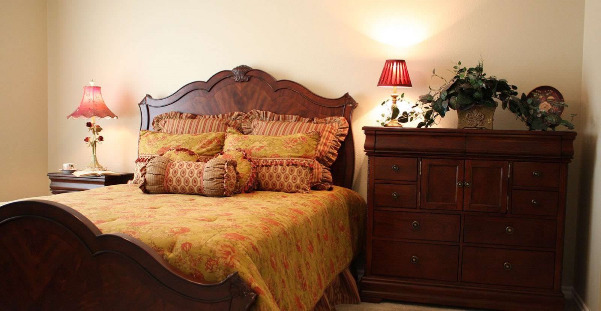 A cozy bedroom featuring a wooden bed with ornate pillows and a matching dresser, softly illuminated by a lamp.