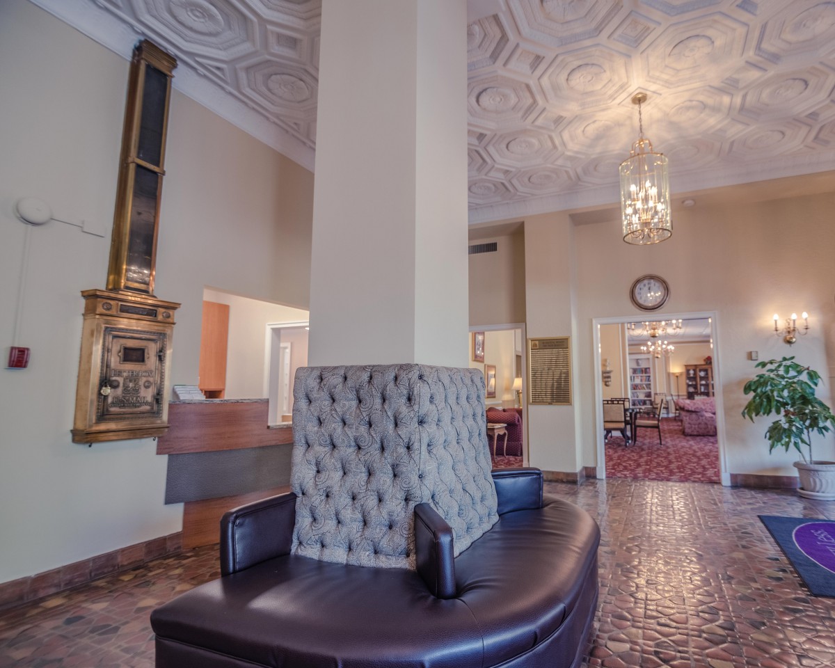 A classic hotel lobby featuring a decorative ceiling and chandelier, with comfortable seating around a central pillar.