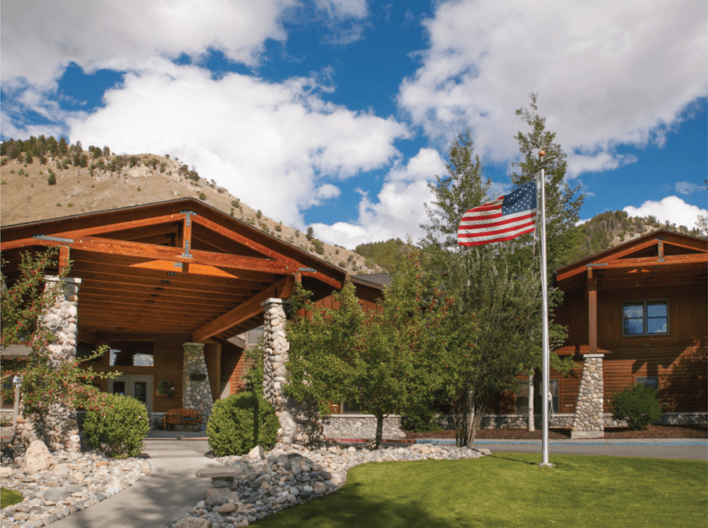 A scenic view of a building entrance with an American flag, surrounded by trees and set against a backdrop of blue sky and clouds.