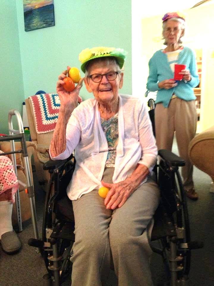 An elderly woman in a wheelchair smiling and holding colorful balls, wearing a bright hat during a celebration.