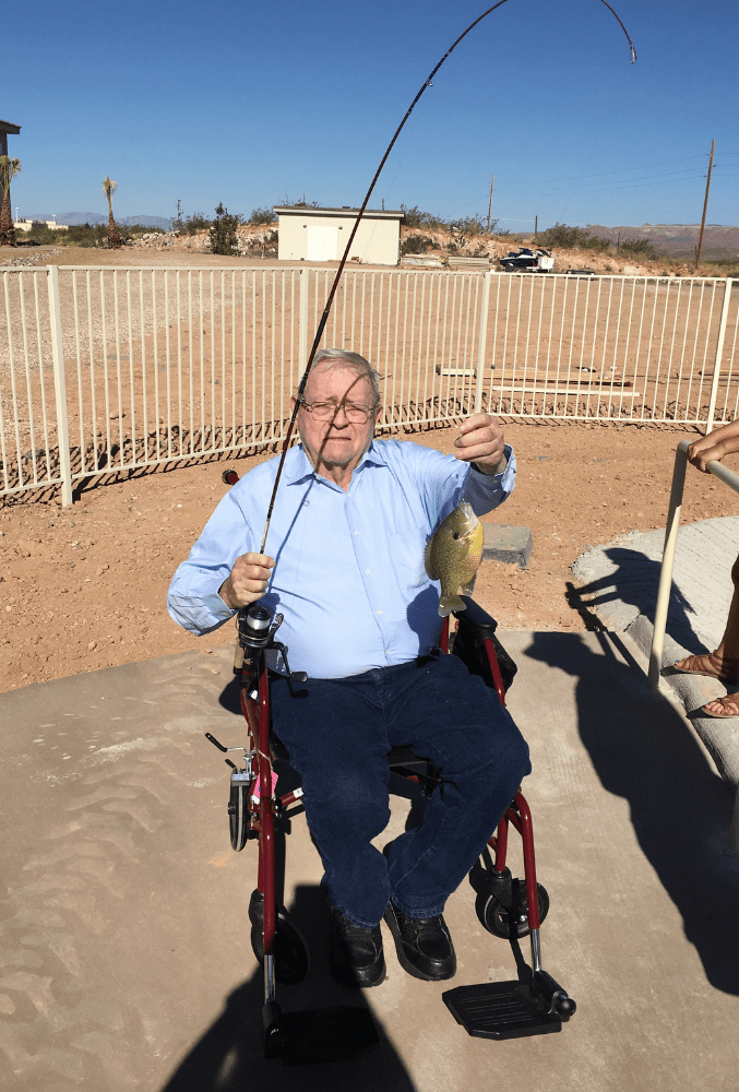A man in a wheelchair holding a fishing rod and a caught fish on a sunny day.