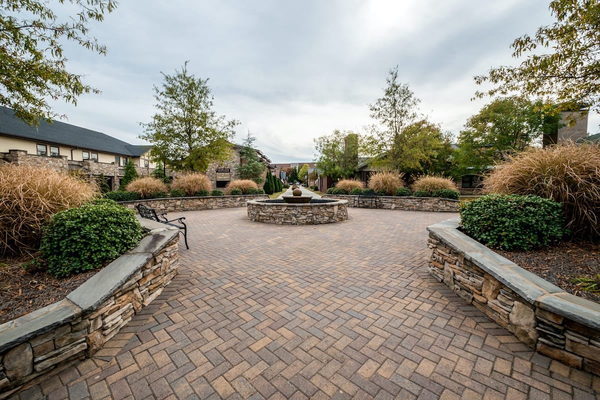 A peaceful courtyard with a central fountain, surrounded by trees and benches on a brick-paved area.