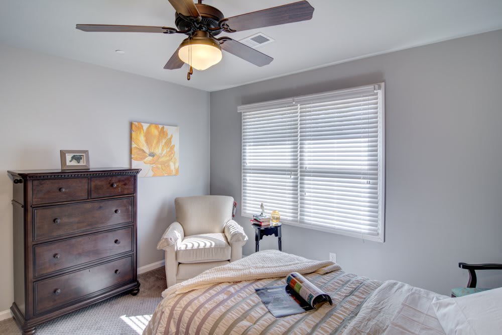 A cozy bedroom with a comfortable armchair, neatly made bed, large window with blinds, and a ceiling fan overhead.