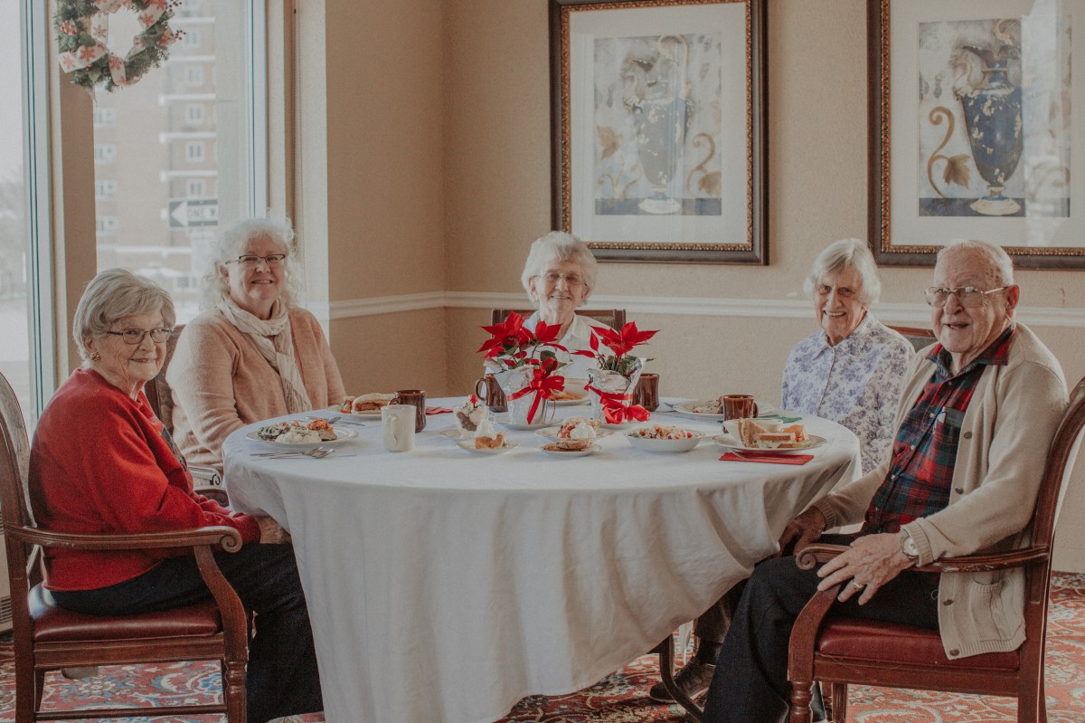 A group of elderly individuals socializing around a table with flowers in an indoor setting.