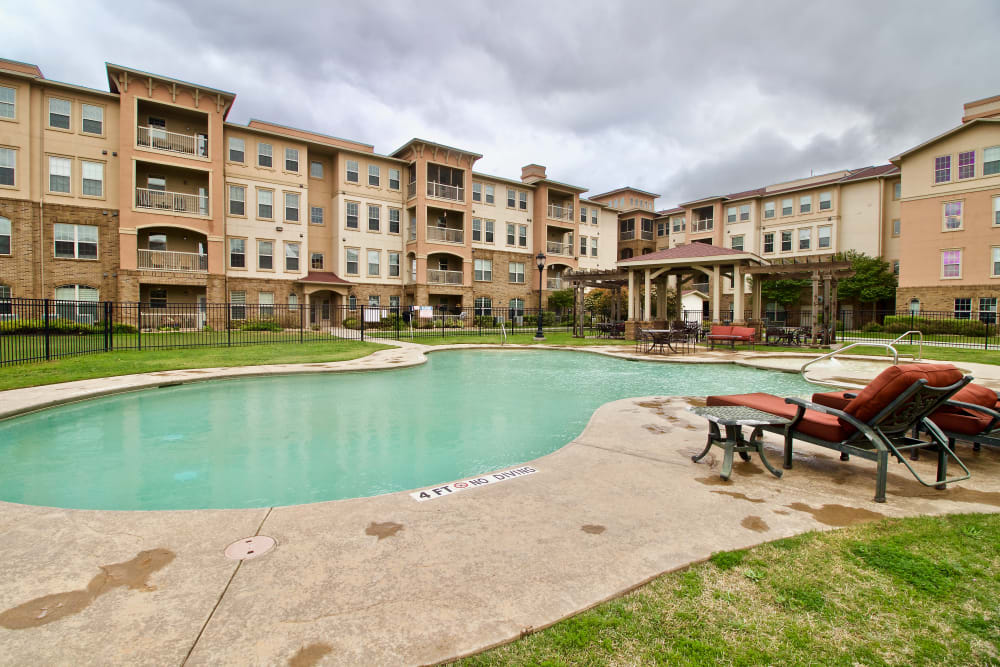 A residential apartment complex with a swimming pool and patio furniture under a cloudy sky.