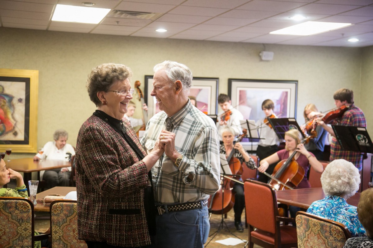 An elderly couple is dancing together while a live orchestra plays in the background.
