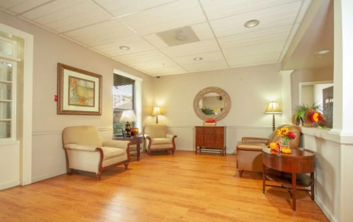 A cozy waiting room featuring comfortable chairs, a table with a lamp, artwork, and a mirror on the wall over a wooden floor.