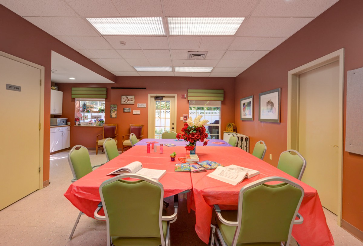 A cozy dining room with a large table covered in a red tablecloth, surrounded by green chairs and decorated with flowers.