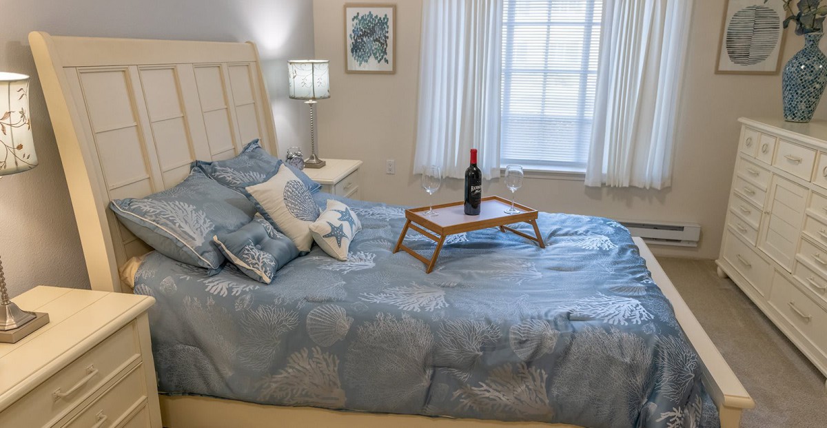 A cozy bedroom with a neatly made bed featuring blue bedding, decorative pillows, and a tray with a wine bottle. A lamp stands on a bedside table next to the window with white curtains.