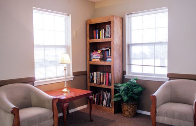 A cozy reading nook with a bookshelf, armchairs, and a warm lamp between two bright windows.