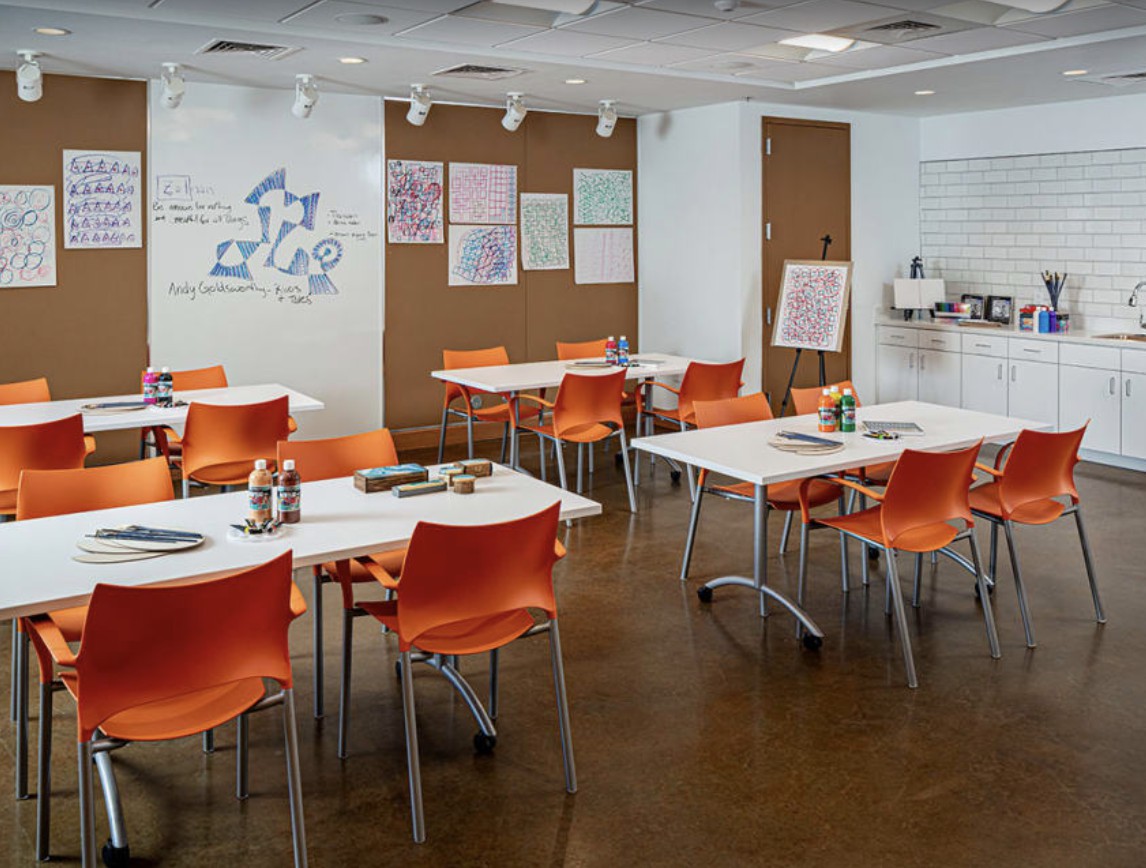 An art classroom with orange chairs and white tables arranged for group activities.