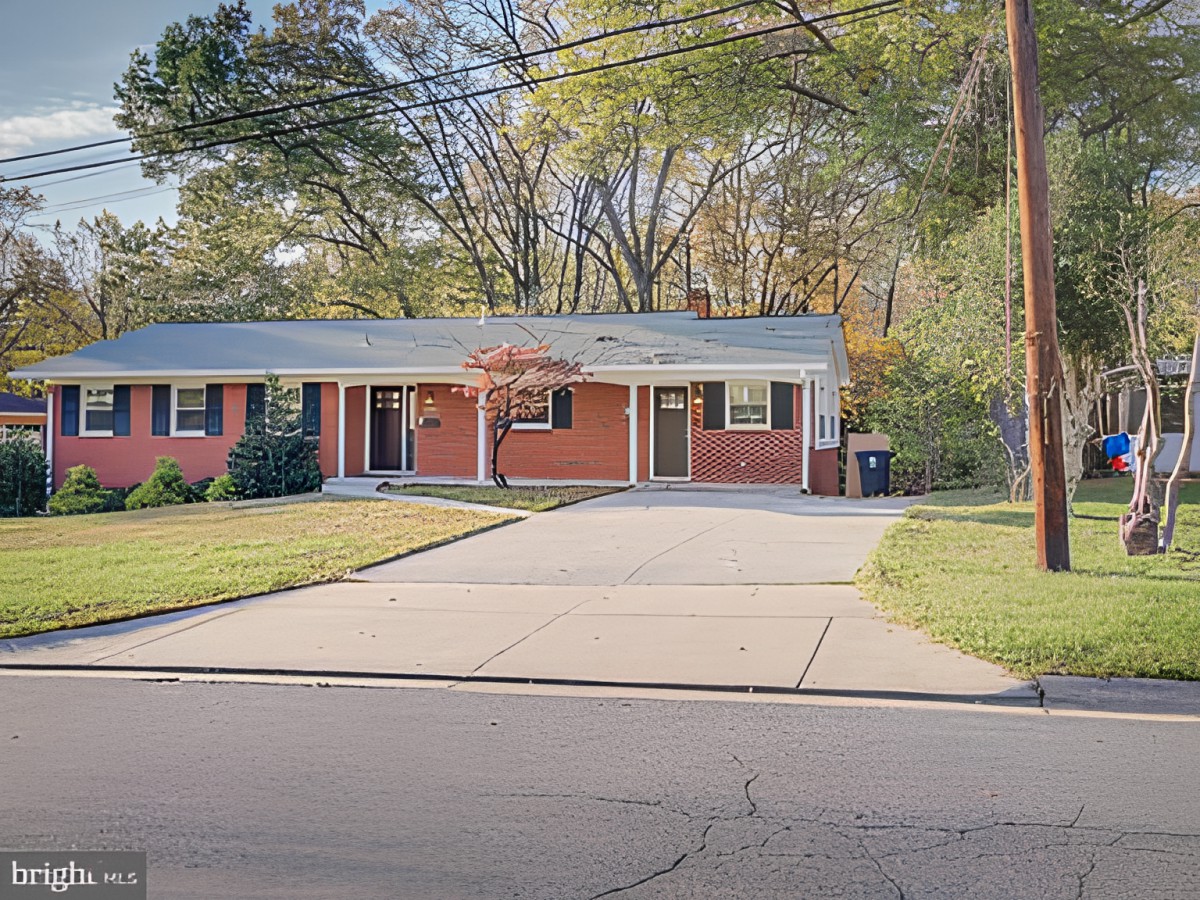 A suburban single-story brick house with a driveway and surrounded by trees.