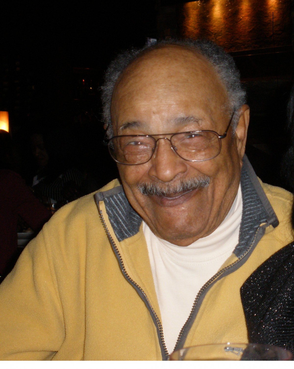 A smiling elderly man wearing glasses and a jacket indoors.
