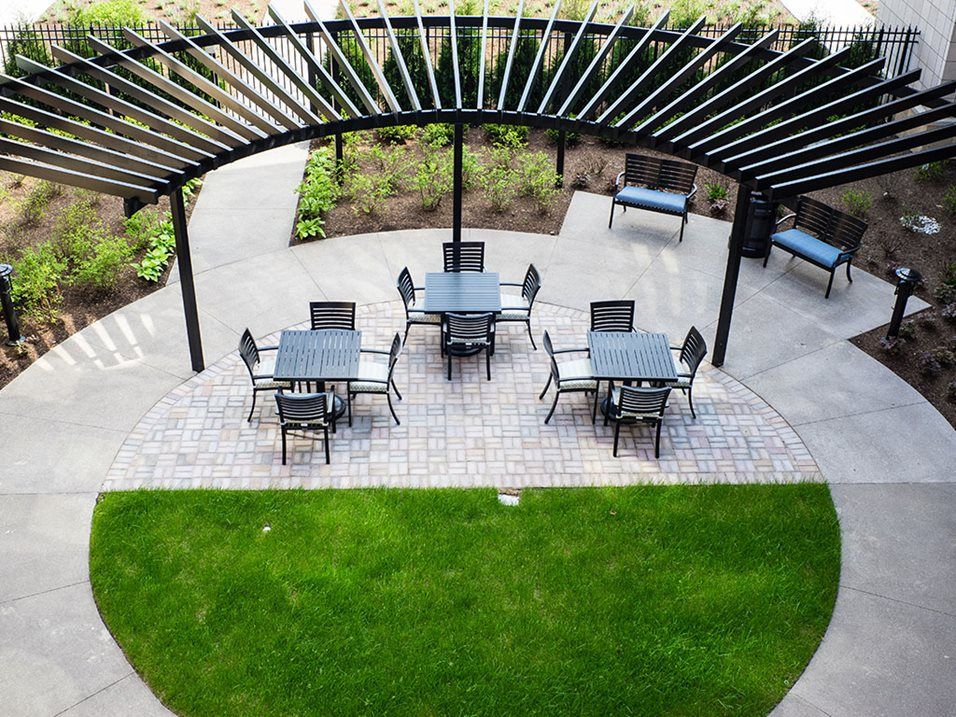 An outdoor patio setup with tables and chairs under a pergola, surrounded by greenery.