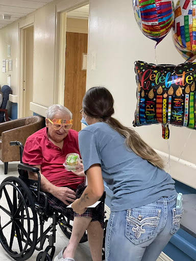 A heartfelt moment of celebration between a caregiver and an elderly individual in a wheelchair, surrounded by festive balloons.