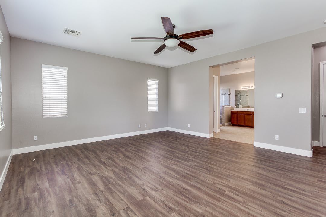 A spacious and empty room with hardwood flooring, featuring a ceiling fan and neutral-colored walls.